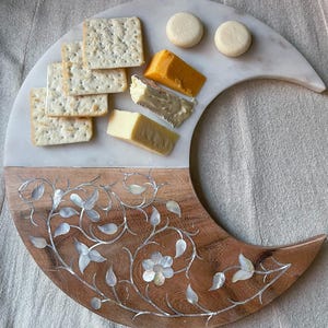 May include: A crescent-shaped serving board with a marble top and wooden base. The wooden portion is decorated with inlaid mother-of-pearl floral designs. The board holds crackers, cheese, and round biscuits, perfect for appetizers.