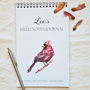 May include: A spiral-bound notebook titled Leo's FIELD NOTES JOURNAL with a watercolor illustration of a red cardinal on a branch. The cover includes the text SIGHTINGS • BIRD SKETCHING • NATURE NOTES and is accompanied by a silver pen.