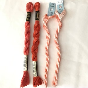 May include: Four spools of embroidery floss, two are a deep orange color and two are a gradient of peach and white. The spools are labeled with the brand name "DMC" and the color number. The spools are also labeled with the brand name "Eavar Co." and the color name "NOVA".