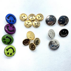 May include: Assortment of smiley face buttons in various colors, including gold, silver, green, purple, and blue. The buttons have a raised smiley face design and the word "smile" written on them.