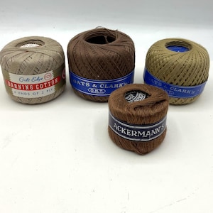 May include: Four spools of thread in various shades of brown and tan. The spools have labels with text including "Gilt Edge Darning Cotton" and "Oats & Clark's O.N.T.". The thread is tightly wound, and the spools are arranged on a white surface.
