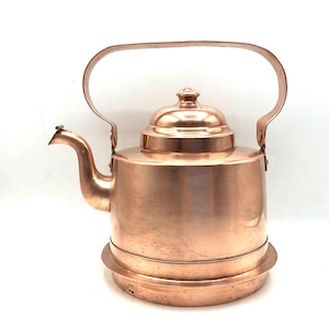 May include: A copper kettle with a curved spout, a domed lid, and a large handle. The kettle sits on a circular base. The copper has a warm, reddish-brown color and a polished finish. A classic kitchenware item.