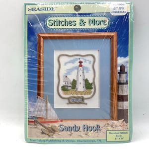 May include: A framed cross-stitch kit featuring a lighthouse scene. The kit includes a wooden frame, a light blue mat, and a stitched image of a white lighthouse with a red top. The finished stitch size is 6" x 8". The kit is titled "Sandy Hook".