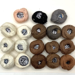May include: A collection of yarn balls in various colors, including pink, black, gray, white, and shades of brown. Each ball has a label with text and a wheat-like design. The yarn appears to be made of wool or a similar fiber, suitable for knitting or crochet projects.