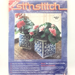 May include: A needlepoint craft kit featuring two blue and white planter designs with pink flowers. The kit includes yarn, a needle, and a yarn threader. The packaging says "Stitnstitch" and "Makes Two".