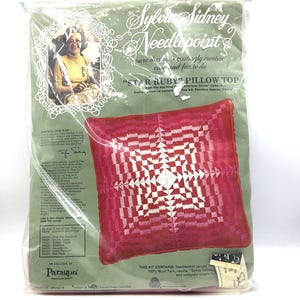 May include: A sealed package containing a needlepoint kit. The kit features a red and white starburst design pillow top. The packaging includes a photo of Sylvia Sidney and text that reads "Star Ruby Pillow Top".