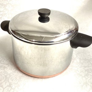 May include: A stainless steel pot with a copper bottom and a black lid with a knob. The pot has two black handles.