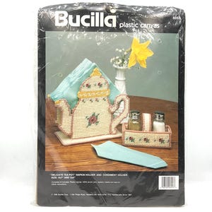 May include: A Bucilla plastic canvas craft kit for a tea pot napkin holder and condiment holder. The packaging displays the finished cream-colored items with pink and green floral accents, measuring 6 inches by 7 inches and 3 inches by 4 inches.