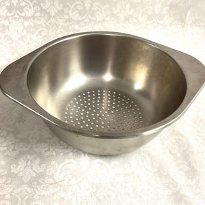 May include: A stainless steel colander with a perforated base and two side handles. The bowl is round with a slightly flared rim. The colander is designed for draining liquids from food items.