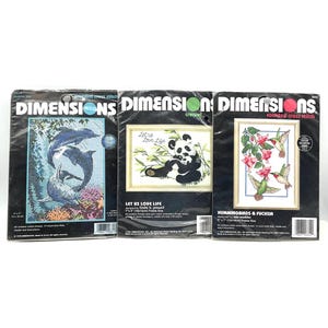 May include: Three Dimensions counted cross stitch kits. The kits feature images of dolphins, a panda bear with the text "Let us Love Life", and hummingbirds with fuchsia flowers. Each kit is in a black package.
