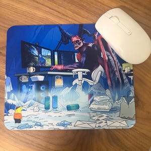 May include: A rectangular mousepad featuring a colorful illustration of a horned demon sitting at a computer desk in a snowy landscape, with a white computer mouse resting on the top right corner of the pad. The scene includes various items.