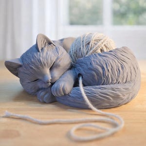 May include: A gray cat-shaped yarn bowl with a sleeping cat design. The bowl holds a ball of light gray yarn, with the yarn strand extending out. The cat is curled up, with its eyes closed, and is resting on a wooden surface.