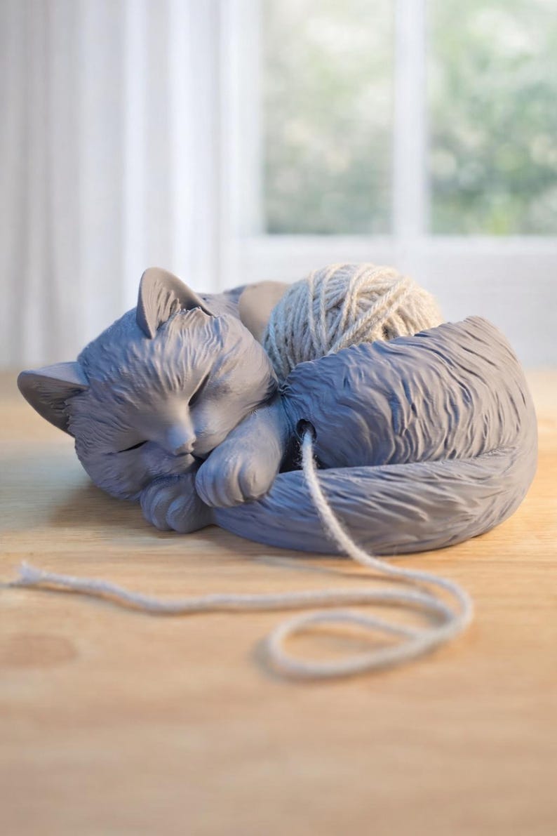 May include: A gray cat-shaped yarn bowl with a sleeping cat design. The bowl holds a ball of light gray yarn, with the yarn strand extending out. The cat is curled up, with its eyes closed, and is resting on a wooden surface.