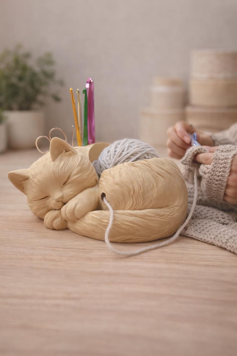 May include: A beige cat-shaped yarn holder with a sleeping cat design. The holder contains a ball of gray yarn and several crochet hooks. A person is crocheting in the background. A charming and functional item for crafters.