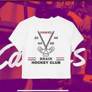 Puede incluir: Una camiseta blanca de manga corta con un gráfico impreso en la espalda que muestra dos palos de hockey cruzados, un disco, el texto HAWKS, 22, 46, 44, 66, BRAIR HOCKEY CLUB y dos líneas horizontales en colores rojo, blanco y azul.