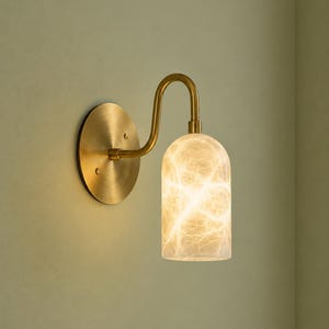 May include: A wall sconce featuring a circular brass backplate and a curved brass arm supporting a cylindrical, translucent glass shade. The shade displays a white, marble-like veined pattern and emits a warm, soft glow from within the fixture.