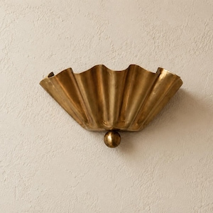 Op de afbeelding: Een messing wandlamp met een brede, waaiervormige kap met diepe, verticale plooien. Het armatuur is aan de onderkant verankerd met een enkel, glad, bolvormig messing detail, wat zorgt voor een gestructureerde en metalen uitstraling.