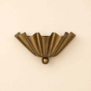 Op de afbeelding: Een wandlamp met een halfronde, geplooide messing kap met een matte afwerking. Het armatuur wordt verankerd door een kleine, centraal geplaatste messing bol aan de basis, wat een waaiervormig silhouet creëert dat uitsteekt vanaf de muur.