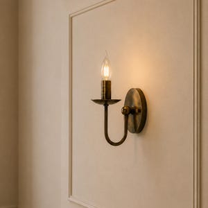 May include: A wall-mounted sconce light fitting with an antique brass finish, featuring a curved arm, a circular backplate, and a single candle-style bulb with a flame-shaped tip that emits a warm, soft glow against the wall surface.