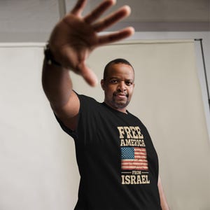 Peut inclure: T-shirt noir avec l'inscription "FREE AMERICA FROM ISRAEL" dans un style vintage. Le motif comprend un drapeau américain avec un effet vieilli. La personne qui porte le t-shirt a la main tendue.
