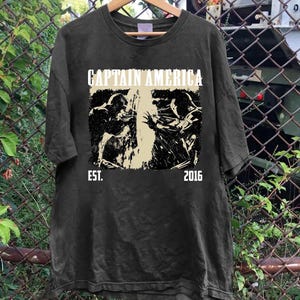 May include: Dark gray t-shirt featuring the text "CAPTAIN AMERICA" above a black and white graphic of Captain America in action. The text "EST. 2016" is below the graphic. The shirt is hanging on a wooden hanger.