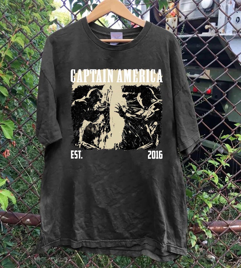 May include: Dark gray t-shirt featuring the text "CAPTAIN AMERICA" above a black and white graphic of Captain America in action. The text "EST. 2016" is below the graphic. The shirt is hanging on a wooden hanger.