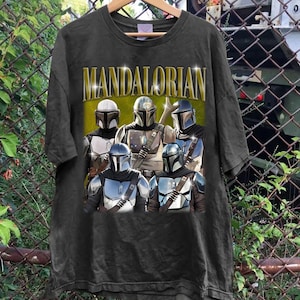 K&ouml;nnte beinhalten: Schwarzes T-Shirt mit einer Gruppe von Mandalorianer-Charakteren in R&uuml;stung. Das Wort "MANDALORIAN" ist in goldenen Buchstaben &uuml;ber den Charakteren aufgedruckt. Das T-Shirt h&auml;ngt an einem Holzb&uuml;gel.