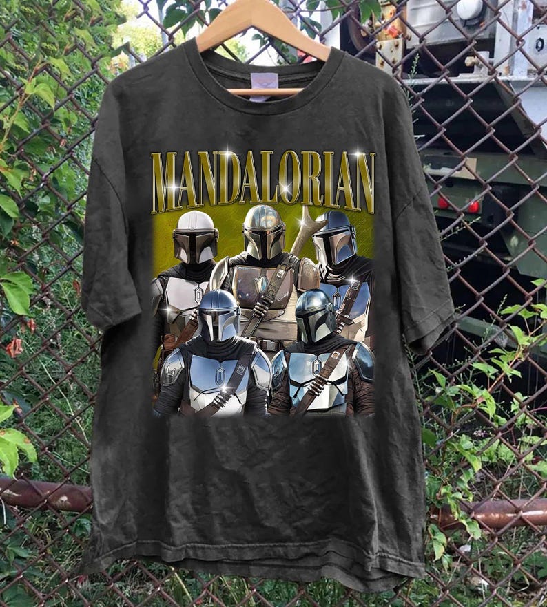 K&ouml;nnte beinhalten: Schwarzes T-Shirt mit einer Gruppe von Mandalorianer-Charakteren in R&uuml;stung. Das Wort "MANDALORIAN" ist in goldenen Buchstaben &uuml;ber den Charakteren aufgedruckt. Das T-Shirt h&auml;ngt an einem Holzb&uuml;gel.