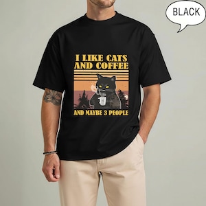 May include: Black t-shirt with a graphic featuring a black cat holding a coffee cup, with the text "I LIKE CATS AND COFFEE AND MAYBE 3 PEOPLE" in yellow and white. The design has a retro sunset and forest background.