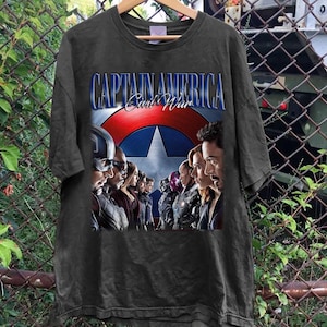 Puede incluir: Camiseta gris oscuro con el texto "CAPTAIN AMERICA Civil War" y un gr&aacute;fico del escudo del Capit&aacute;n Am&eacute;rica y varios personajes. La camiseta est&aacute; colgada en una percha de madera.
