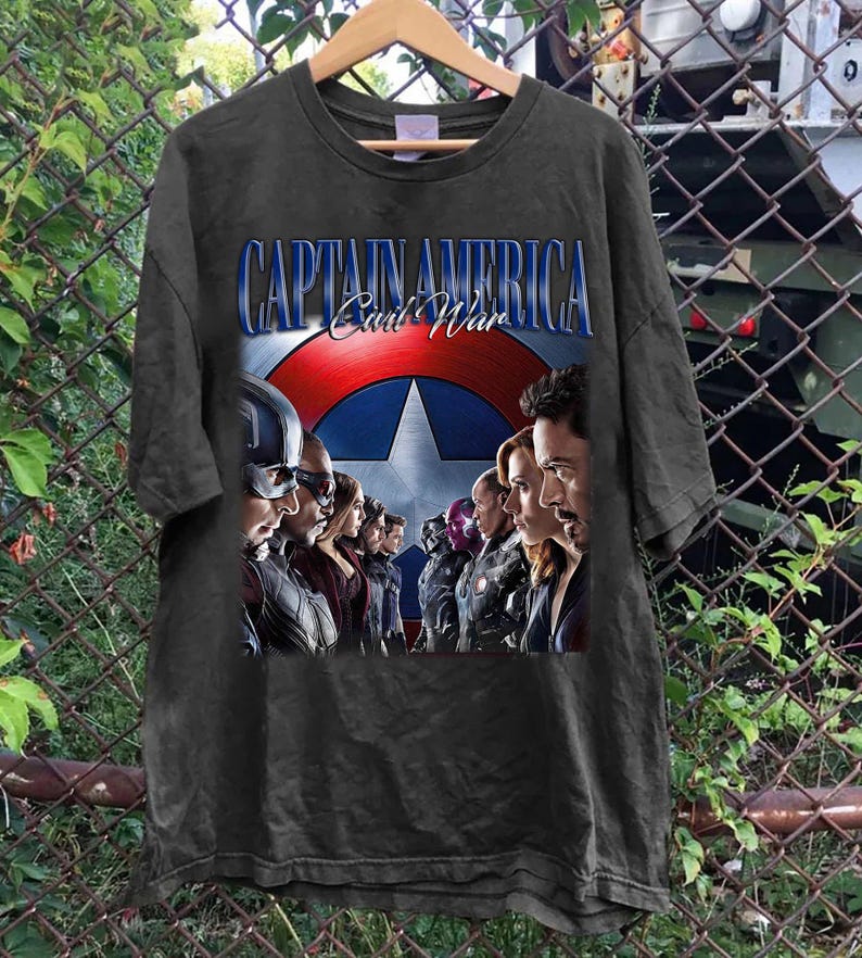 Puede incluir: Camiseta gris oscuro con el texto "CAPTAIN AMERICA Civil War" y un gr&aacute;fico del escudo del Capit&aacute;n Am&eacute;rica y varios personajes. La camiseta est&aacute; colgada en una percha de madera.