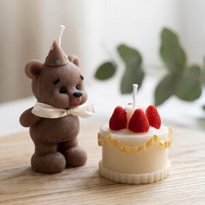 May include: A brown teddy bear candle wearing a party hat and a white bow, next to a small white cake candle topped with red strawberry candles. The candles are on a wooden surface.
