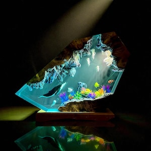 May include: A unique, illuminated resin sculpture depicting an underwater scene. The artwork features a whale, colorful coral, fish, and jellyfish, all encased in a clear, glowing resin. The sculpture is mounted on a wooden base.