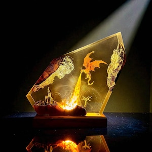 Puede incluir: Escultura de resina de temática fantástica que representa un dragón naranja lanzando fuego hacia un castillo. La obra de arte está encerrada en un bloque de resina transparente y angular con detalles de pan de oro y se asienta sobre una base de madera. Un foco ilumina la escena.