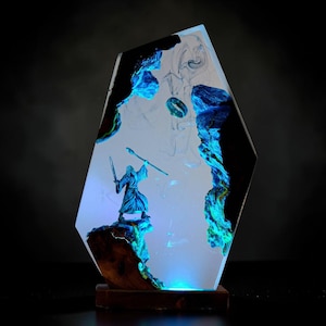 May include: A unique resin sculpture featuring a figure with a staff and sword, set within a glowing blue and white landscape. The sculpture is encased in a clear, geometric form, mounted on a wooden base. The artwork has a fantasy theme.