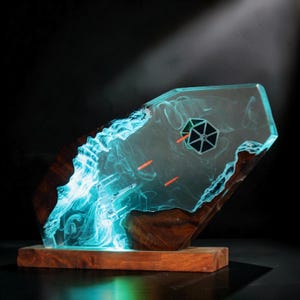 May include: A decorative display piece featuring a translucent blue resin slab embedded with a wooden frame. Inside the resin, a miniature starfighter and a TIE fighter are suspended, with orange laser beams and swirling light effects.