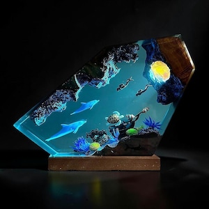 Könnte beinhalten: Ein einzigartiges Kunstwerk aus Harz, das eine Unterwasserszene mit Delfinen, Tauchern und einer Schildkröte darstellt. Das Kunstwerk ist in klarem, blau getöntem Harz mit einem Holzsockel eingeschlossen. Die Szene umfasst einen leuchtenden Mond und Korallen.