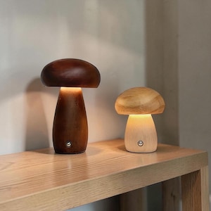 May include: Two mushroom-shaped lamps made of wood. The larger lamp is dark brown, and the smaller lamp is light tan. Both lamps feature a smooth, rounded cap and a cylindrical base with a small, circular silver power button on the front.