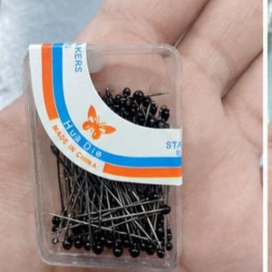 Könnte beinhalten: Ein rechteckiger Behälter aus durchsichtigem Kunststoff, gefüllt mit Stecknadeln mit kleinen schwarzen kugelförmigen Köpfen. Das Etikett zeigt ein orangefarbenes Schmetterlingssymbol, den Text "Hua Die", "MADE IN CHINA" und "STAINLESS" sowie orange und blaue Streifen.