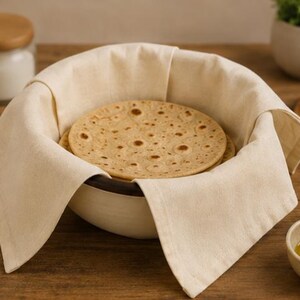 May include: A round, off-white enamel bowl with a dark rim, lined with a plain, light beige fabric cloth. Inside the bowl sits a stack of round, flat, golden-brown toasted bread discs with visible dark brown spots on their surfaces.