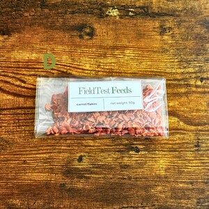 Puede incluir: Una bolsa de pl&aacute;stico transparente que contiene copos de zanahoria, con una etiqueta que dice "Field Test Feeds". La etiqueta tambi&eacute;n indica "copos de zanahoria" y un peso neto de 30g. La bolsa est&aacute; sobre una superficie de madera.