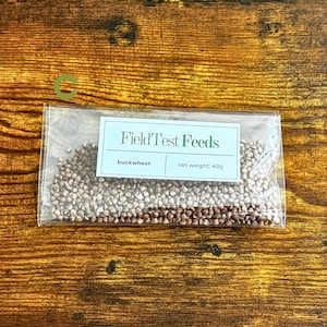 Puede incluir: Un paquete transparente lleno de trigo sarraceno sobre una superficie de madera. El paquete tiene una etiqueta que dice "FieldTest Feeds" con el texto "trigo sarraceno" y "peso neto: 40g". La superficie de madera es de color marr&oacute;n c&aacute;lido.