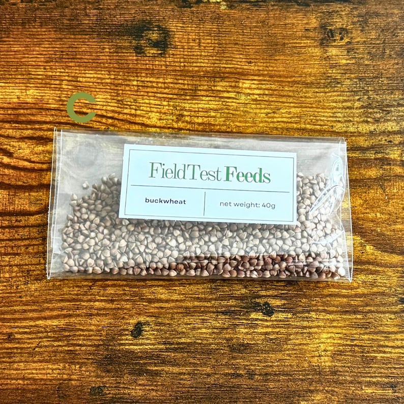 Puede incluir: Un paquete transparente lleno de trigo sarraceno sobre una superficie de madera. El paquete tiene una etiqueta que dice "FieldTest Feeds" con el texto "trigo sarraceno" y "peso neto: 40g". La superficie de madera es de color marr&oacute;n c&aacute;lido.