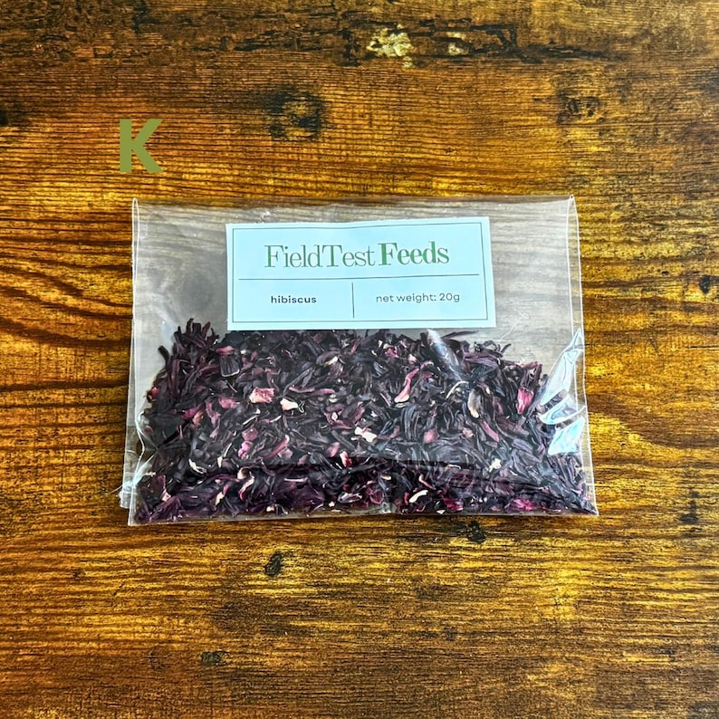 Puede incluir: Una bolsa transparente llena de flores de hibisco secas. La etiqueta dice "FieldTest Feeds" con la palabra "hibiscus" y "peso neto: 20g". Las flores secas son de color rojo intenso.