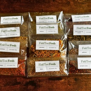 Puede incluir: Una colecci&oacute;n de bolsas de pl&aacute;stico transparente llenas de varios ingredientes secos, cada una etiquetada como "Field Test Feeds". Los ingredientes incluyen gusanos de la harina, avena entera, trigo sarraceno, copos, farro, cal&eacute;ndula y c&aacute;rtamo. Los pesos netos se indican en gramos.