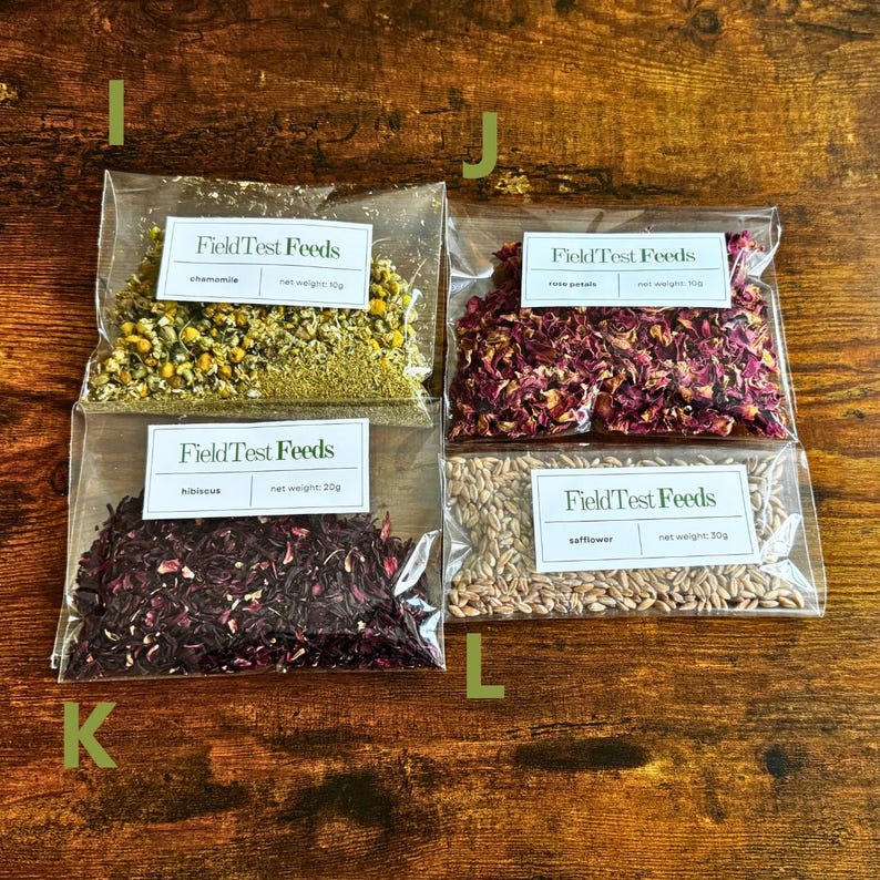 Puede incluir: Cuatro bolsas transparentes de hierbas y flores secas sobre una superficie de madera. Las bolsas contienen manzanilla, p&eacute;talos de rosa, hibisco y c&aacute;rtamo. Cada bolsa tiene una etiqueta blanca con el texto "Field Test Feeds". Pesos netos: 10g, 20g y 30g.