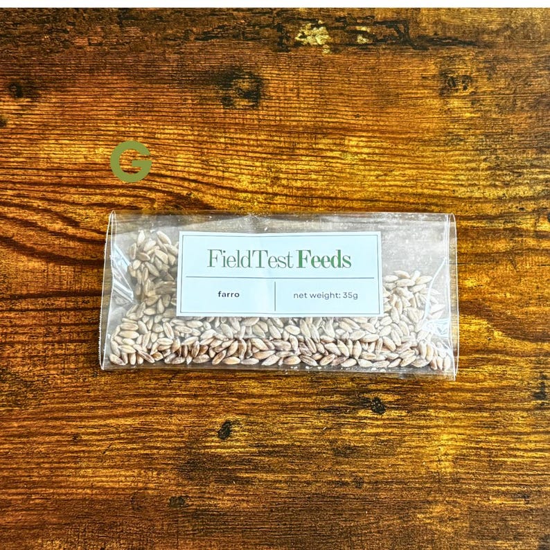 Puede incluir: Una bolsa transparente llena de granos de farro, con una etiqueta que dice "FieldTest Feeds". La etiqueta tambi&eacute;n indica un peso neto de 35g. La bolsa est&aacute; sobre un fondo de madera.