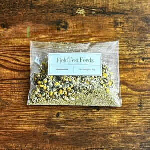 Puede incluir: Una bolsa de pl&aacute;stico transparente llena de flores y hojas de manzanilla secas. La bolsa tiene una etiqueta rectangular con el texto "FieldTest Feeds" y las palabras "manzanilla" y "peso neto: 10g". La bolsa est&aacute; sobre una superficie de madera.