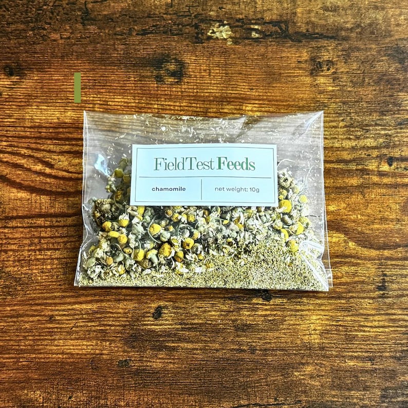 Puede incluir: Una bolsa de pl&aacute;stico transparente llena de flores y hojas de manzanilla secas. La bolsa tiene una etiqueta rectangular con el texto "FieldTest Feeds" y las palabras "manzanilla" y "peso neto: 10g". La bolsa est&aacute; sobre una superficie de madera.