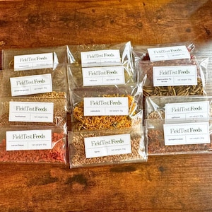 Puede incluir: Una colecci&oacute;n de bolsas de pl&aacute;stico transparente llenas de varios ingredientes secos, cada una etiquetada con "Field Test Feeds". Los ingredientes incluyen granos de avena entera, trigo sarraceno, copos de zanahoria y cal&eacute;ndula. Las bolsas est&aacute;n dispuestas sobre una superficie de madera.